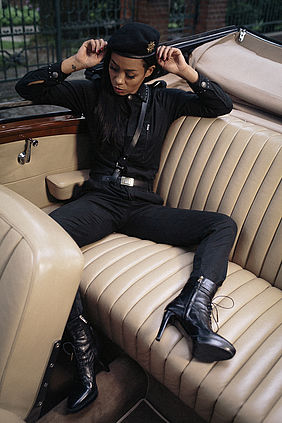 Jadu sitzt schräg und breitbeinig auf der Rücksitzbank eines luxuriösen Cabrios