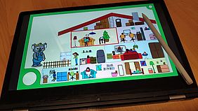Ein Tablet mit einem Stift darauf. Auf dem Bildschirm ein buntes Bild von einem gezeichneten Haus. Ein Koala zeigt auf das Haus.